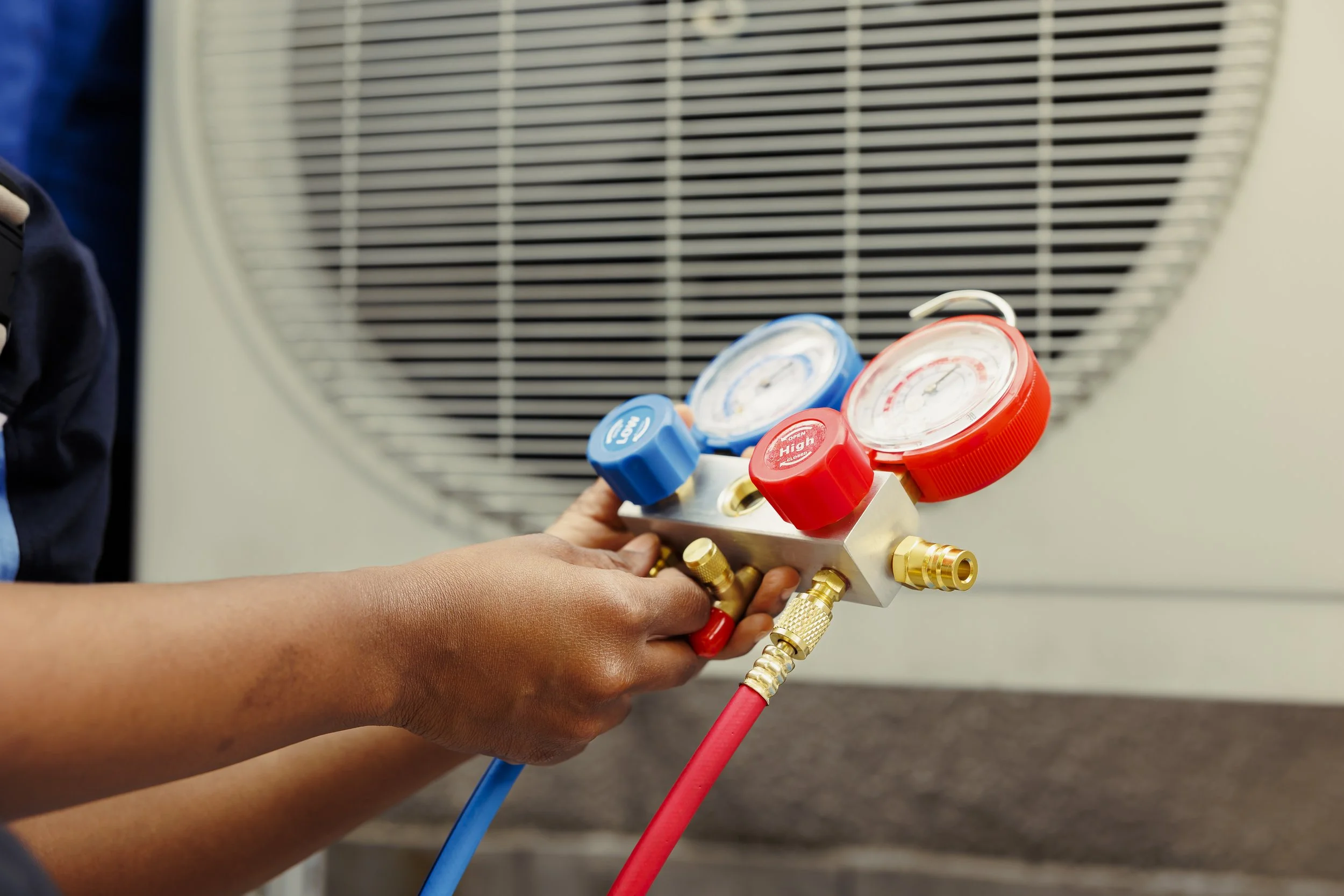 A person holding a refrigeration manifold gauge set with red and blue hoses, used for HVAC refrigeration work, in front of an air conditioning unit.