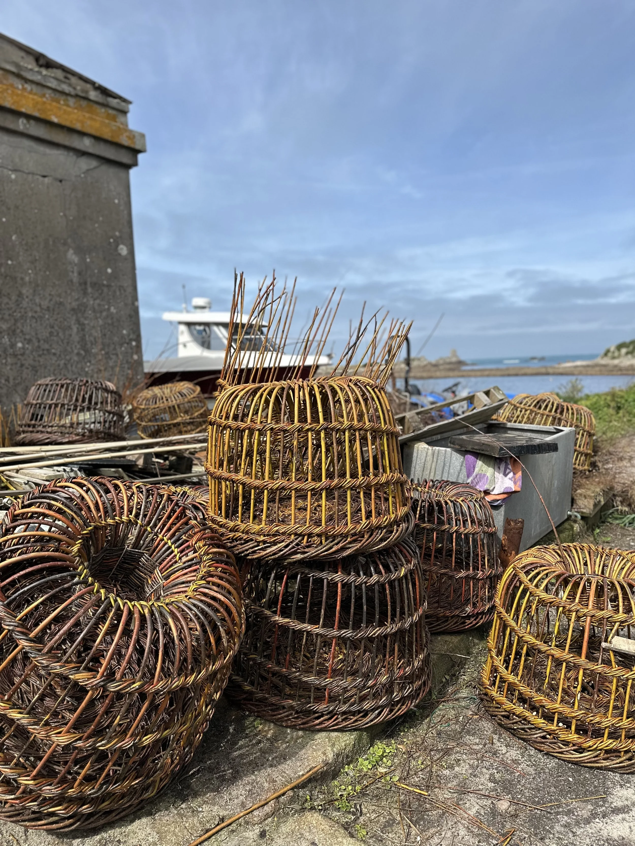 Stacked wicker lobster traps on a concrete surface in St Agnes near the coast with a boat in the background and a cloudy-blue sky.