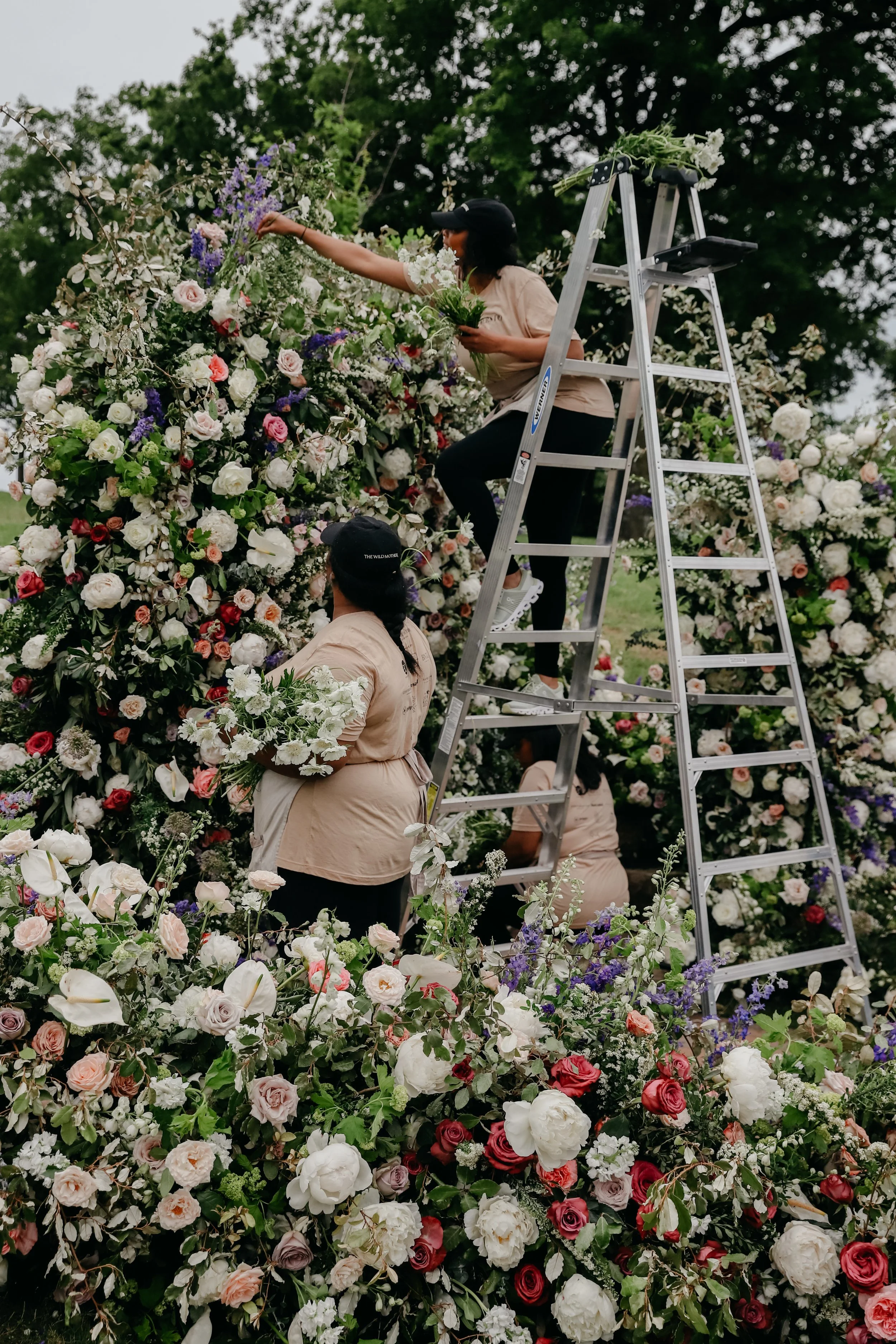 Several women in beige uniforms arranging and attaching flowers on a large floral arch, with one woman on a ladder and the others holding flowers.