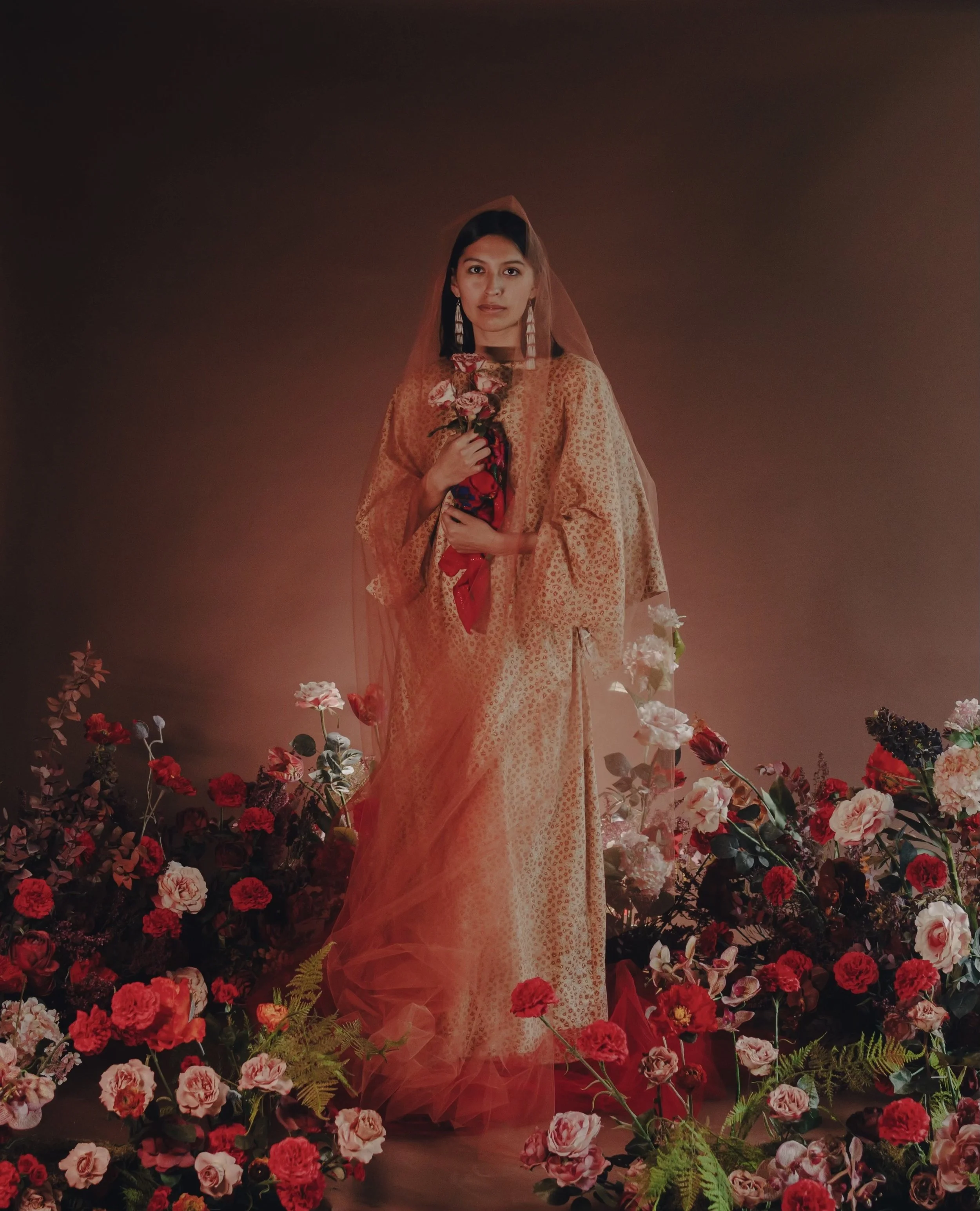 A woman dressed in a gold traditional dress, standing among colorful flowers, holding a bouquet of pink roses, with a brown background.