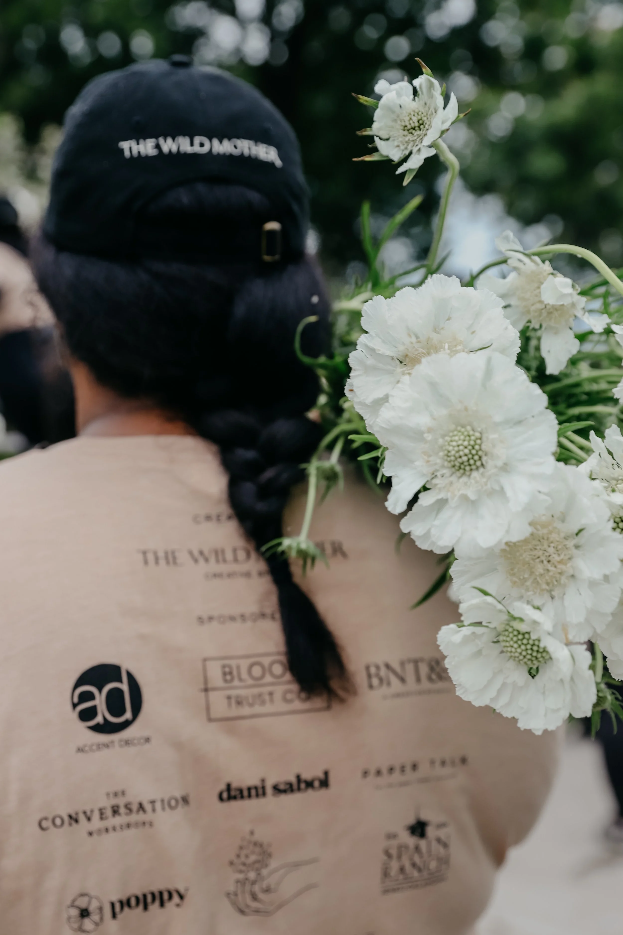 Person with black braided hair wearing a black cap and a beige shirt with various logos, carrying a large bouquet of white flowers.