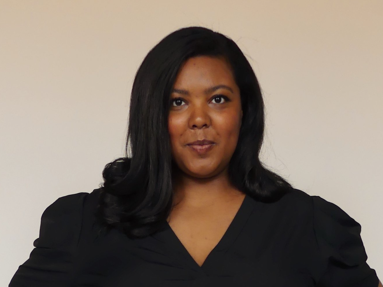 Portrait of a woman with medium-length black hair, wearing a black top, standing against a plain beige background.