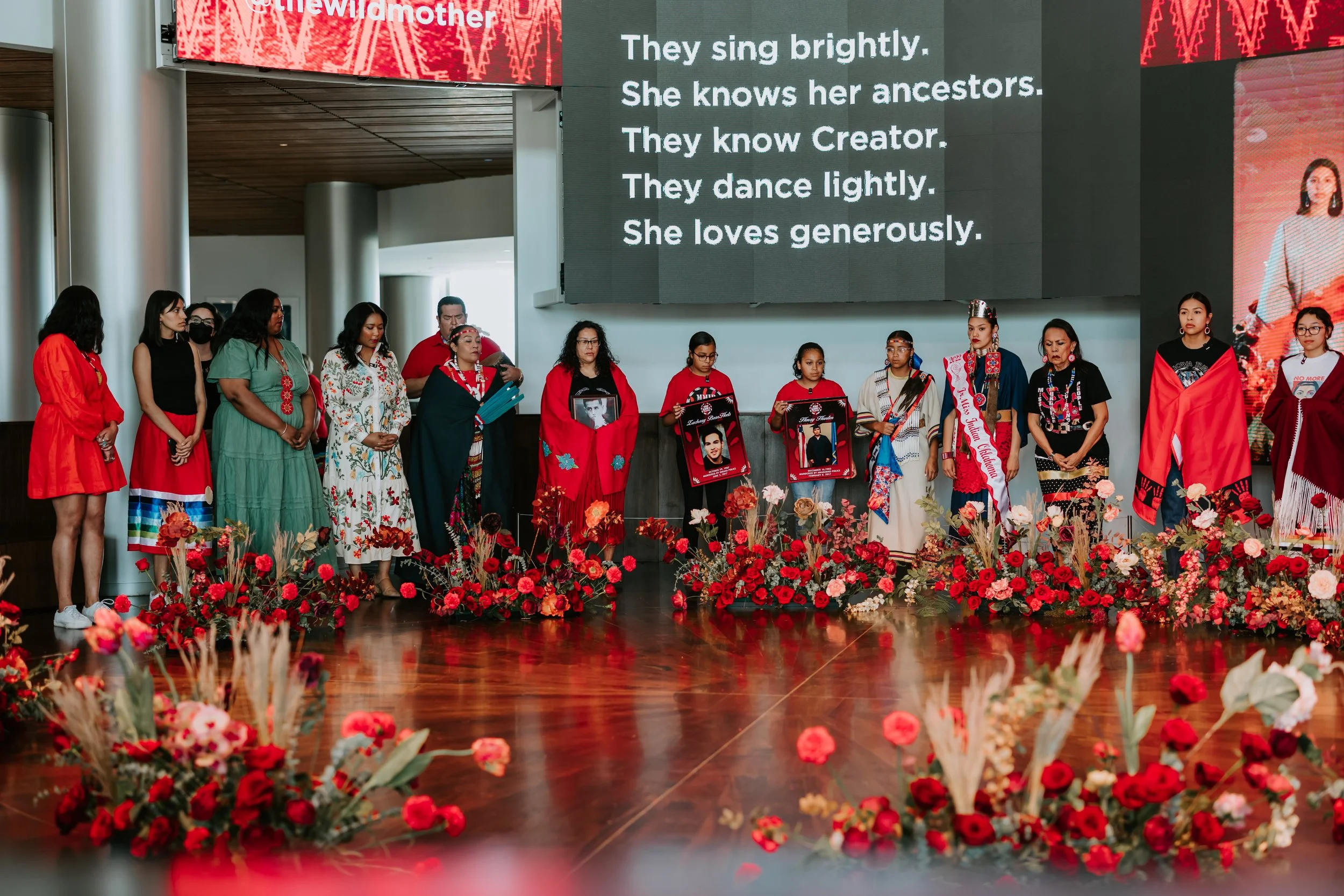 A group of people standing on a stage decorated with red flowers, participating in a ceremony or memorial. Some individuals are holding photos of a young man. Behind them, a screen displays a song lyrics about ancestors, Creator, and love.