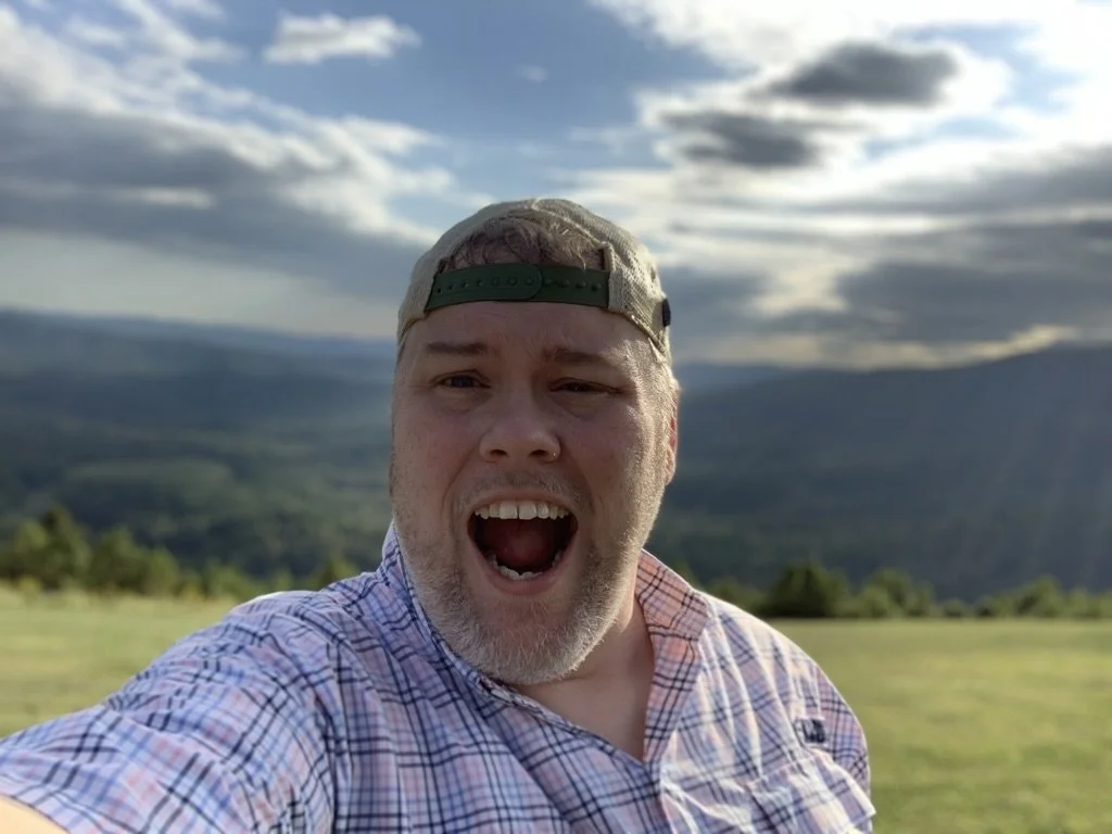 A man with a beard and wearing a baseball cap backwards, taking a selfie outdoors on a sunny day. Background shows green fields and distant mountains under partly cloudy sky.