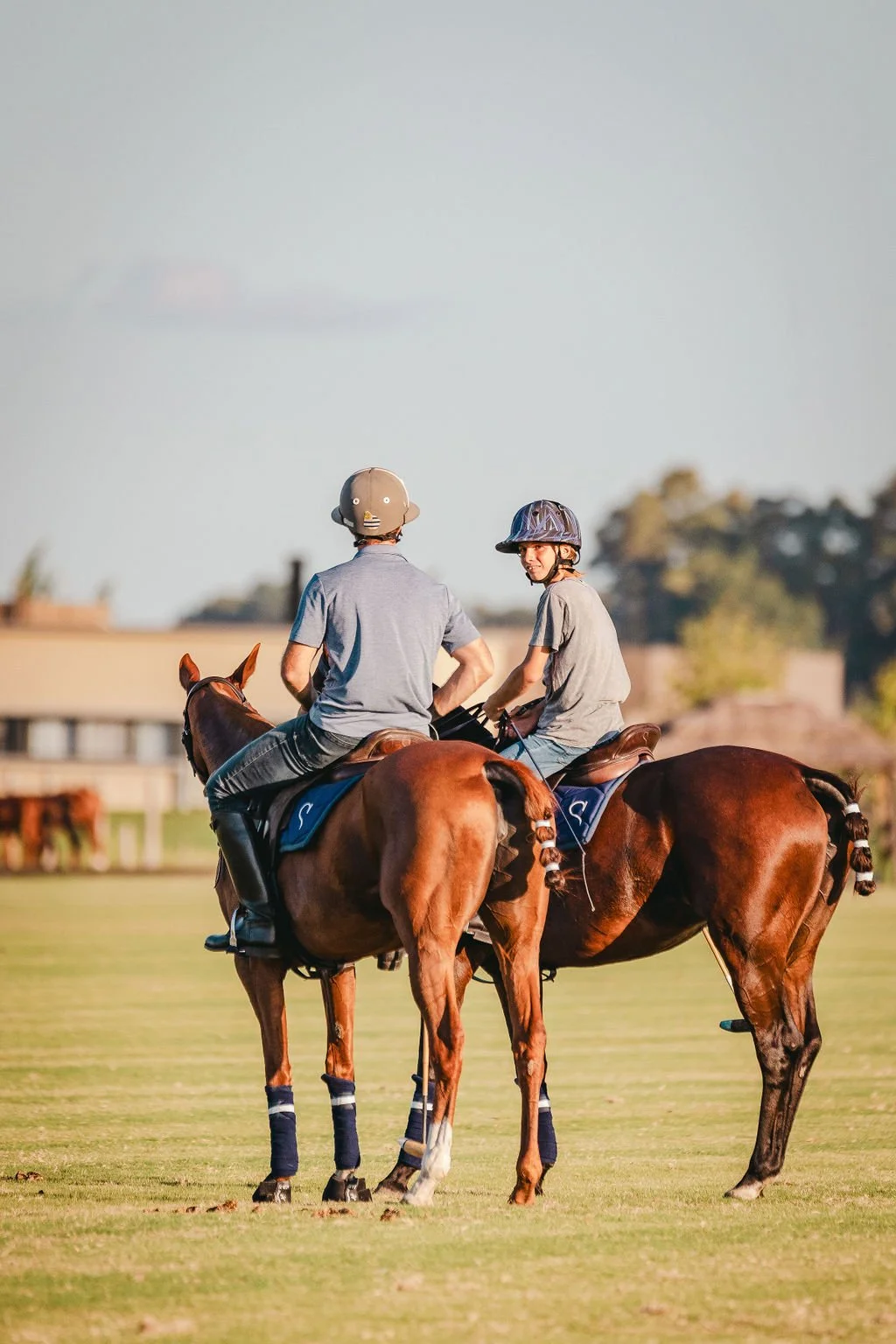A polo player riding a horse on a polo field, seen from behind, wearing a green helmet, blue shirt, and white pants, holding a mallet.