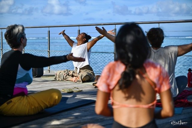 Group of people practicing yoga outdoors on a deck by the water, with a woman leading a pose and others sitting on yoga mats.