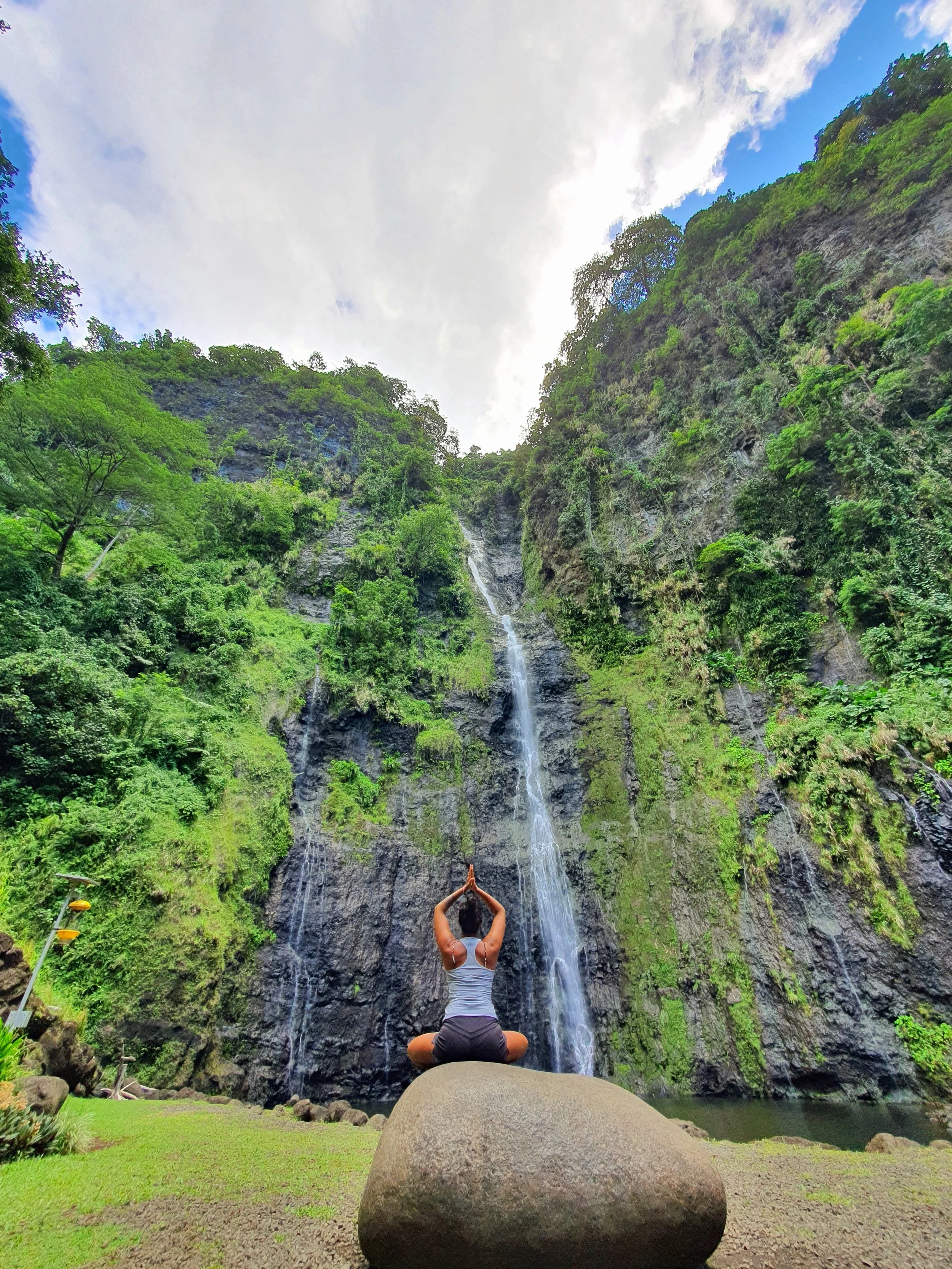 A person practicing yoga in a seated meditation pose on a large rock in front of a tall waterfall surrounded by lush greenery.