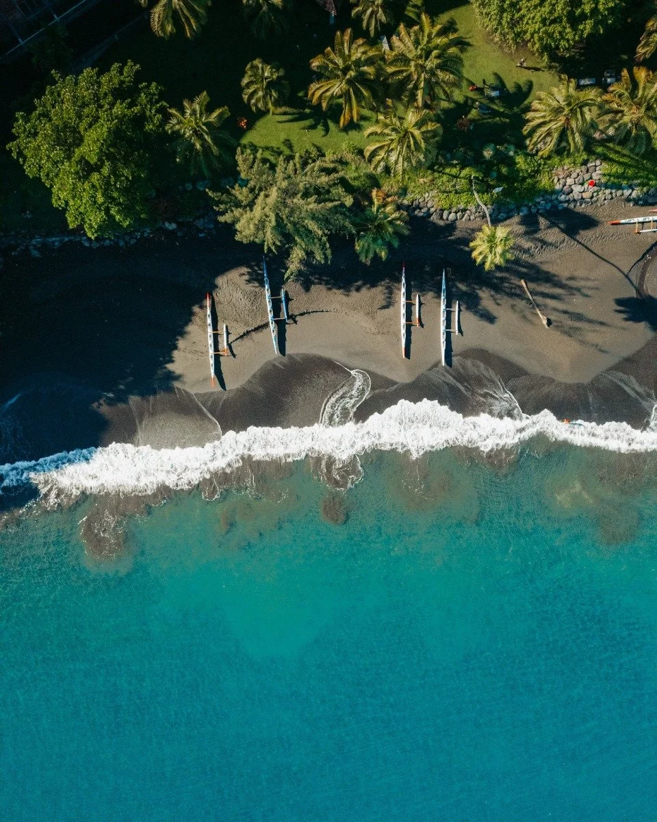 Aerial view of a beach with five paddleboards/ocean kayaks lined up on the sand near the shoreline, with lush greenery and palm trees in the background.