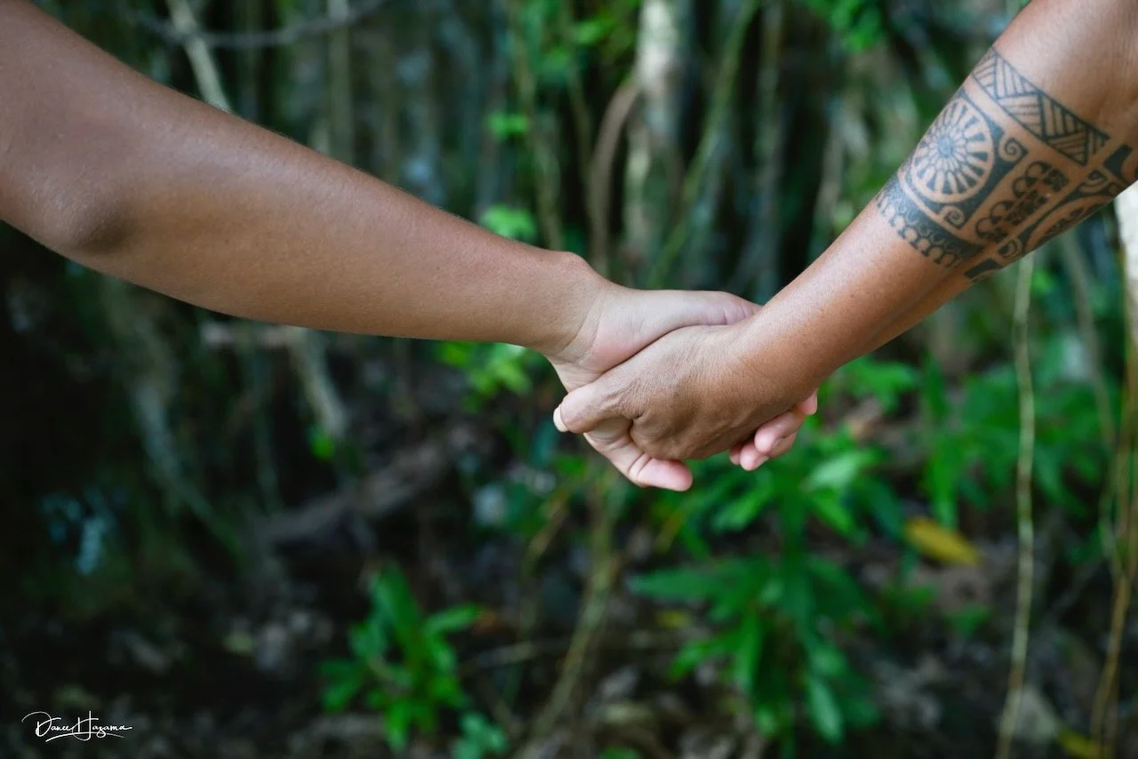 Two people holding hands outdoors in a jungle setting, one with a tattooed arm.