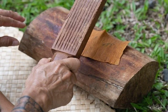 A person carving a piece of wood with a chisel and wooden mallet outdoors, with greenery in the background.