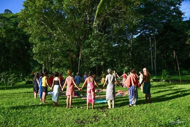 Group of people holding hands in a circle outdoors in a forested area.