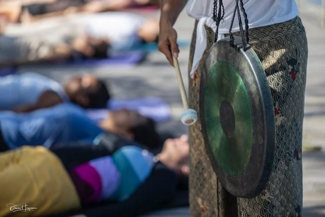 A person holding a gong and a mallet, with a group of people lying on the ground in the background.