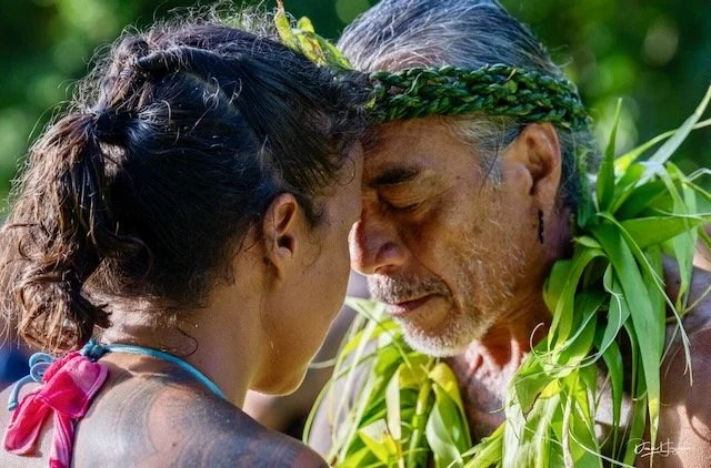 A young woman and an older man with gray hair and a beard are touching foreheads, both with eyes closed, in a face-to-face moment outdoors. The man is wearing a leafy wreath on his head and a garland of green leaves around his shoulders, while the woman has her hair tied back.
