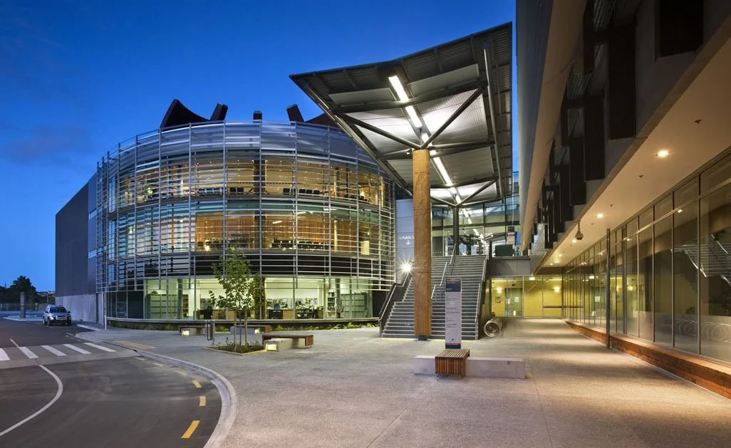Modern building with glass facade and metal grid, illuminated at dusk, with a pedestrian walkway and stairs outside.