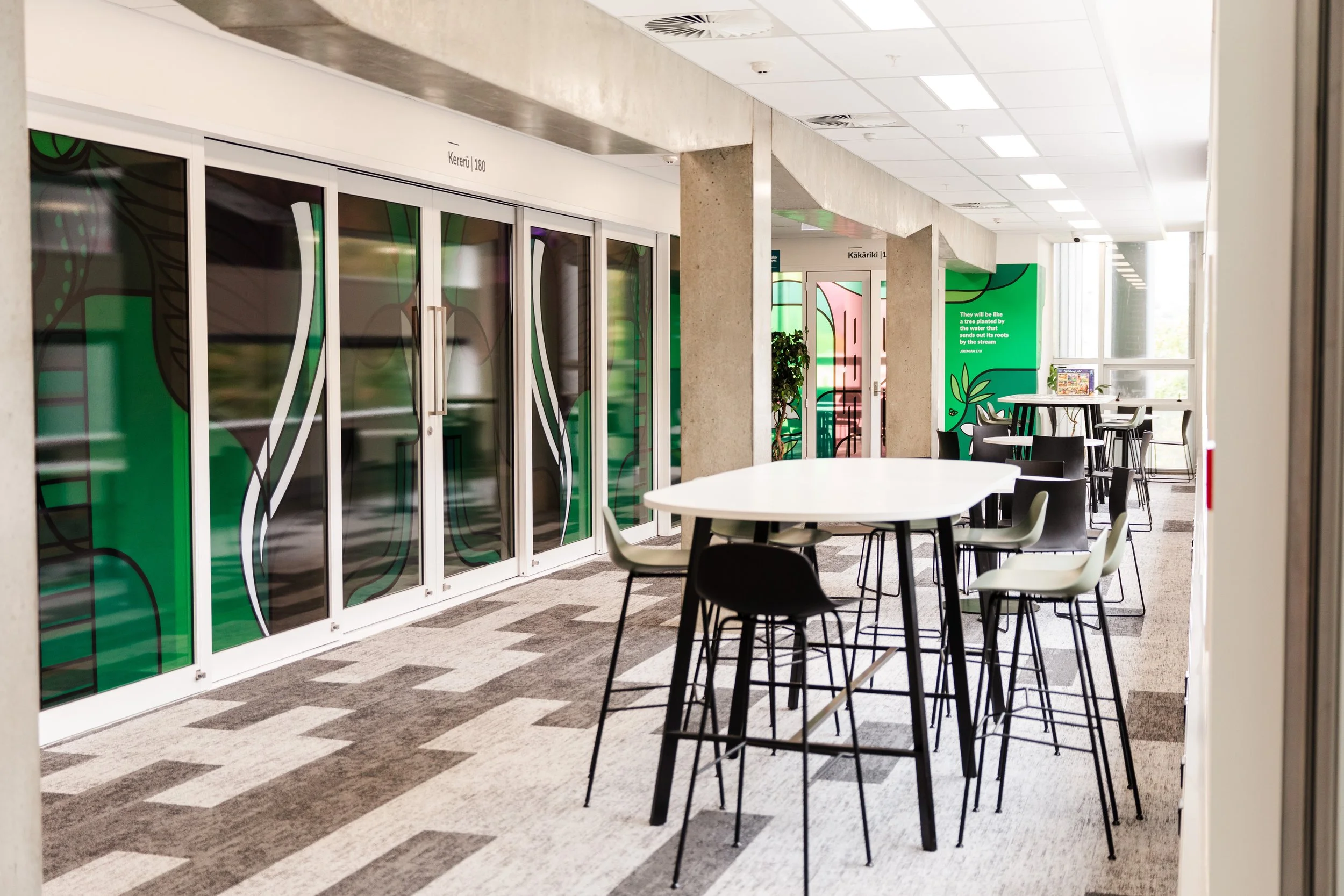 Interior of a modern workspace with high tables, chairs, and glass doors decorated with green and black graphic designs.