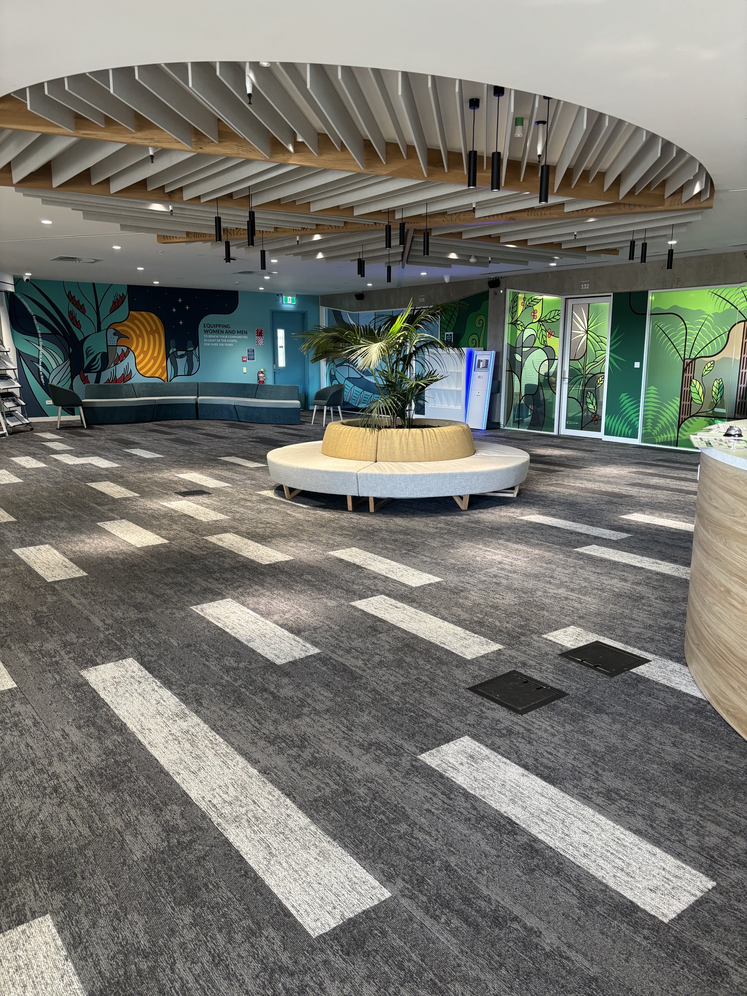 Modern interior space with a patterned carpet, a central circular seating area with plants, colorful wall murals with foliage and animals, and a vending machine.