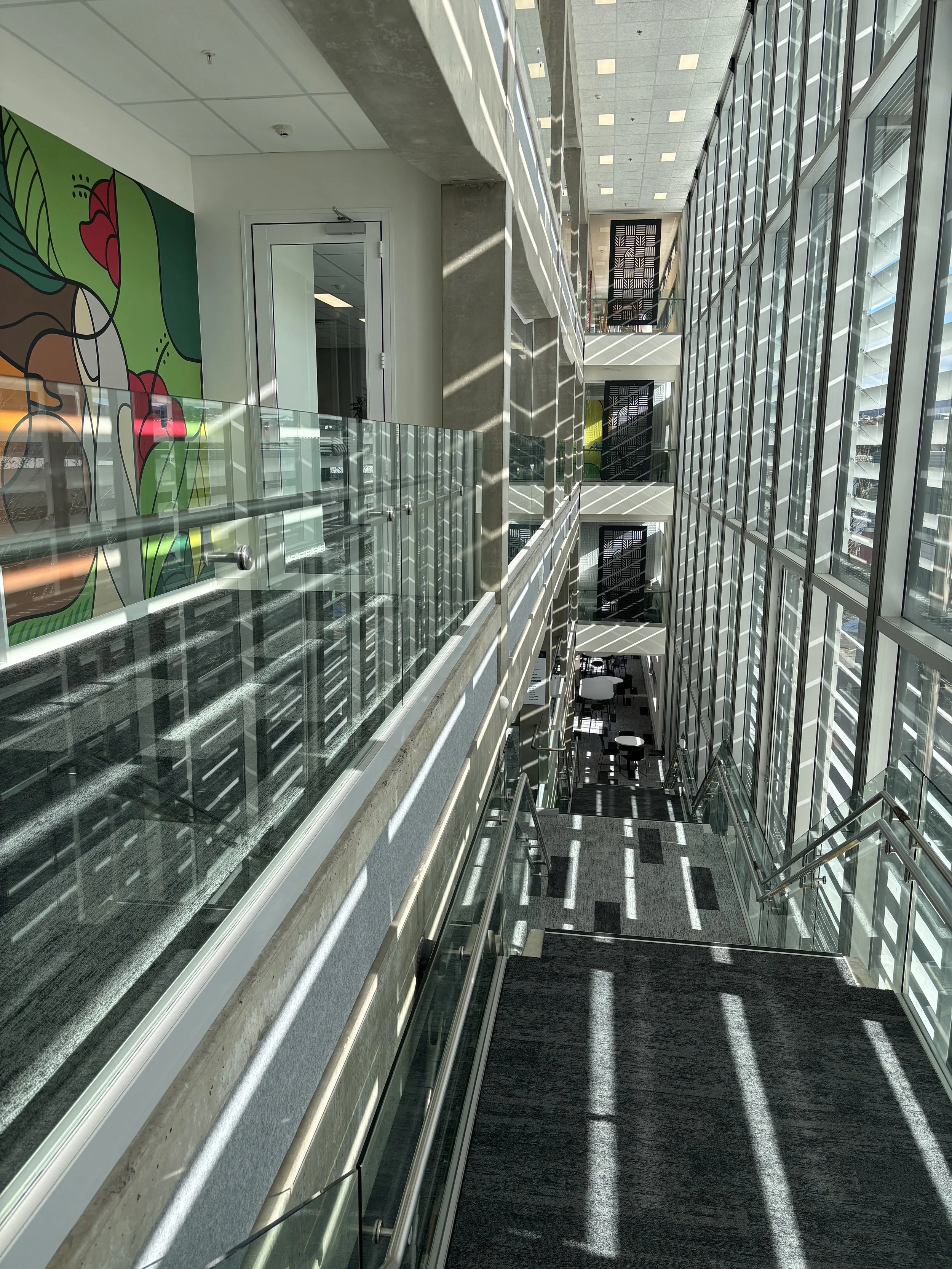 Interior view of a modern building with large glass windows, stairs, and sunlight casting shadows. There is a colorful abstract mural on the wall and a glass railing along the corridor.