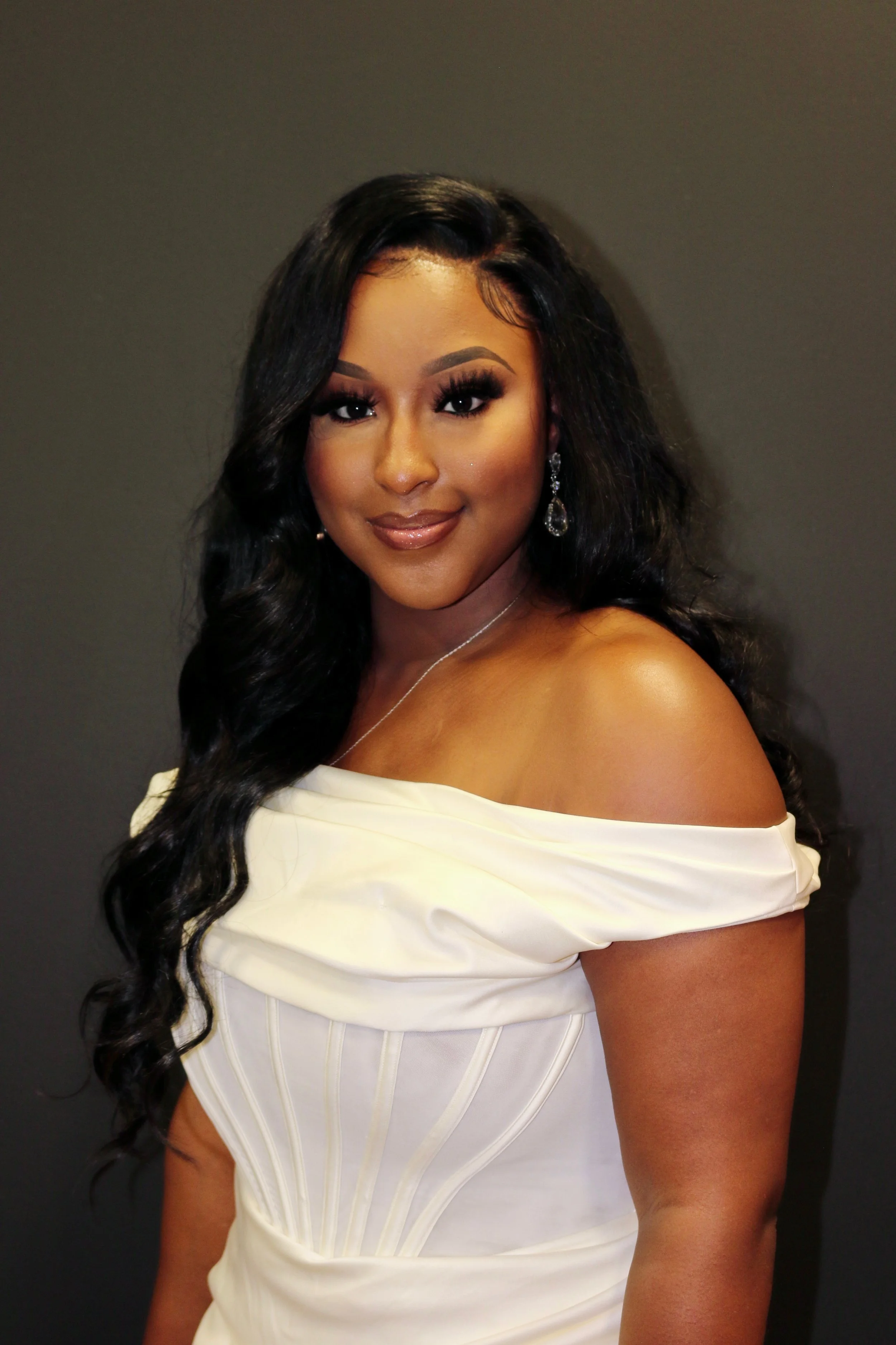 A woman with long black wavy hair wearing a white off-the-shoulder dress and dangling earrings, smiling at the camera against a dark gray background.