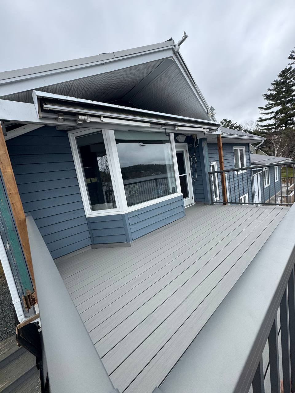 A large outdoor balcony with gray wood decking attached to a blue house with white trim, with a dark railing along the edge and trees in the background.
