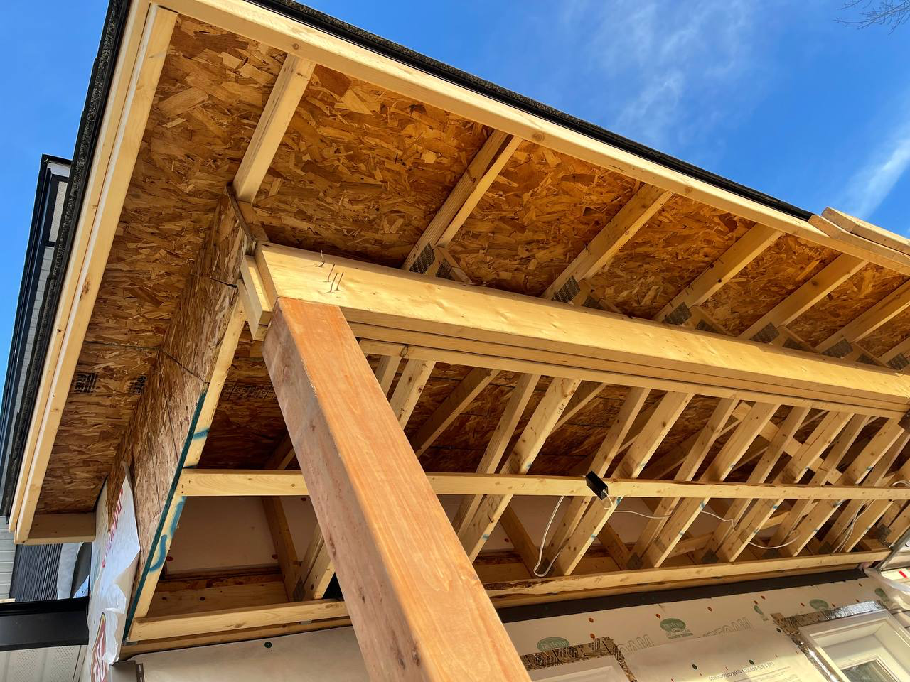 Construction site showing the wooden framework of a building's second floor with exposed OSB sheathing and a beam, against a background of a blue sky.