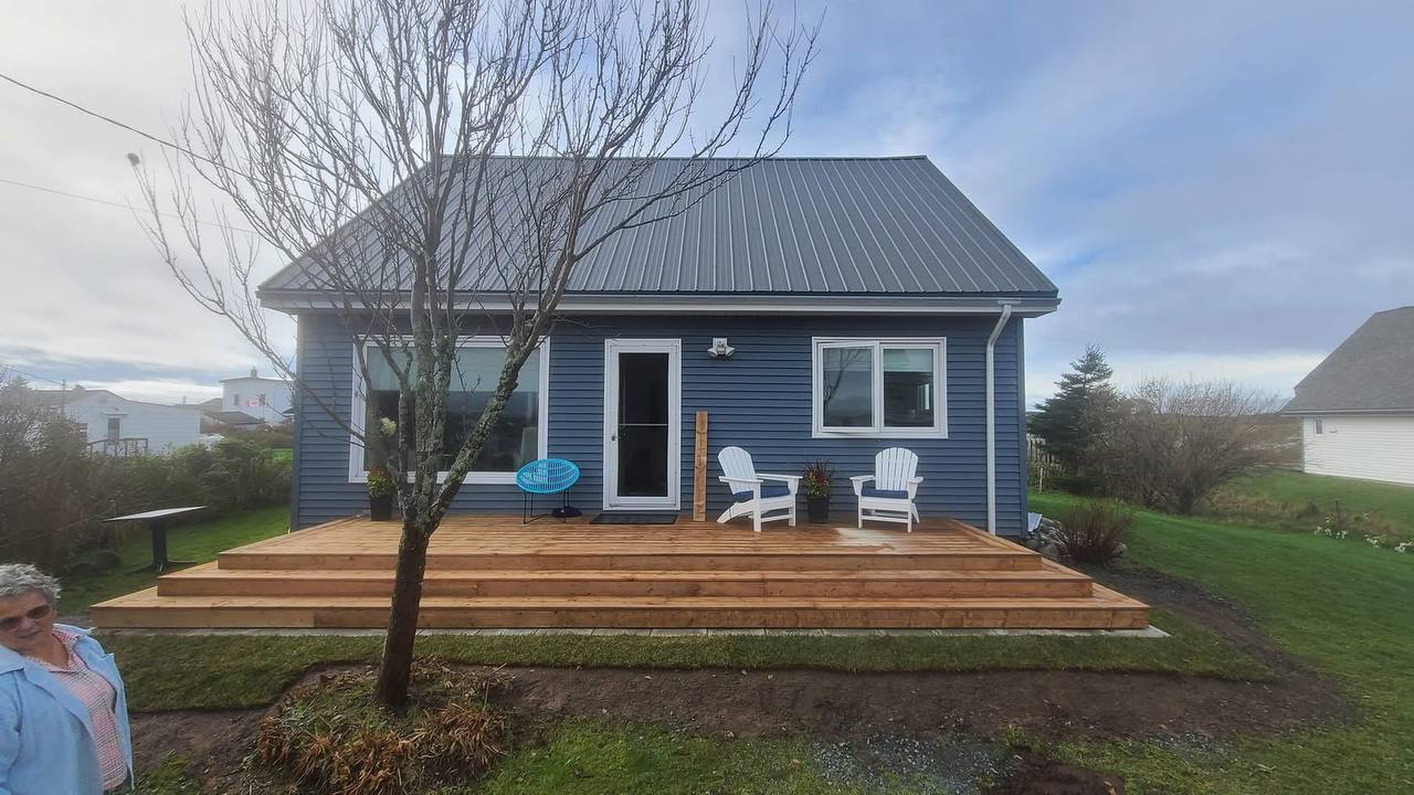 Backyard view of a blue house with a new wooden deck. Two white Adirondack chairs and a blue chair are on the deck. There is a tree in the foreground and grassy yard around.
