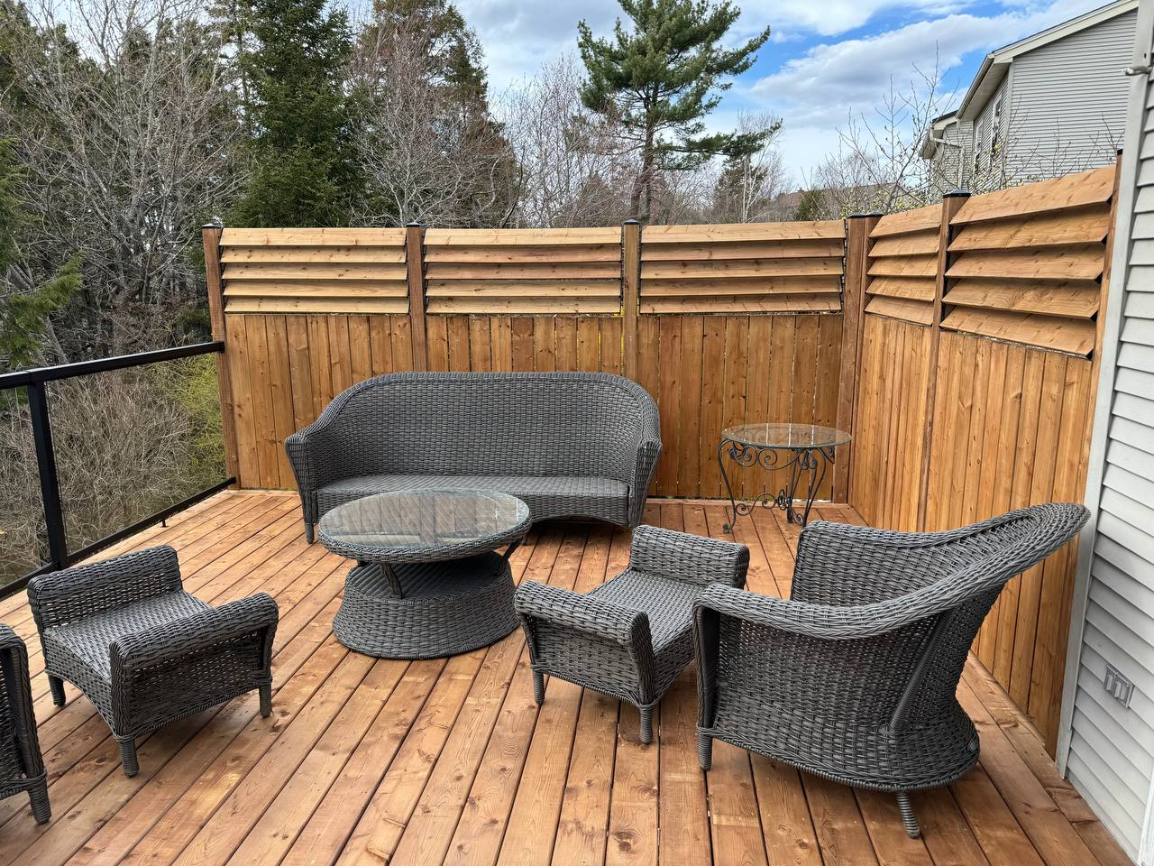 Outdoor wooden deck with wicker furniture, including two armchairs, a loveseat, a coffee table, and two side tables, surrounded by a wooden privacy fence, with trees and a cloudy sky in the background.