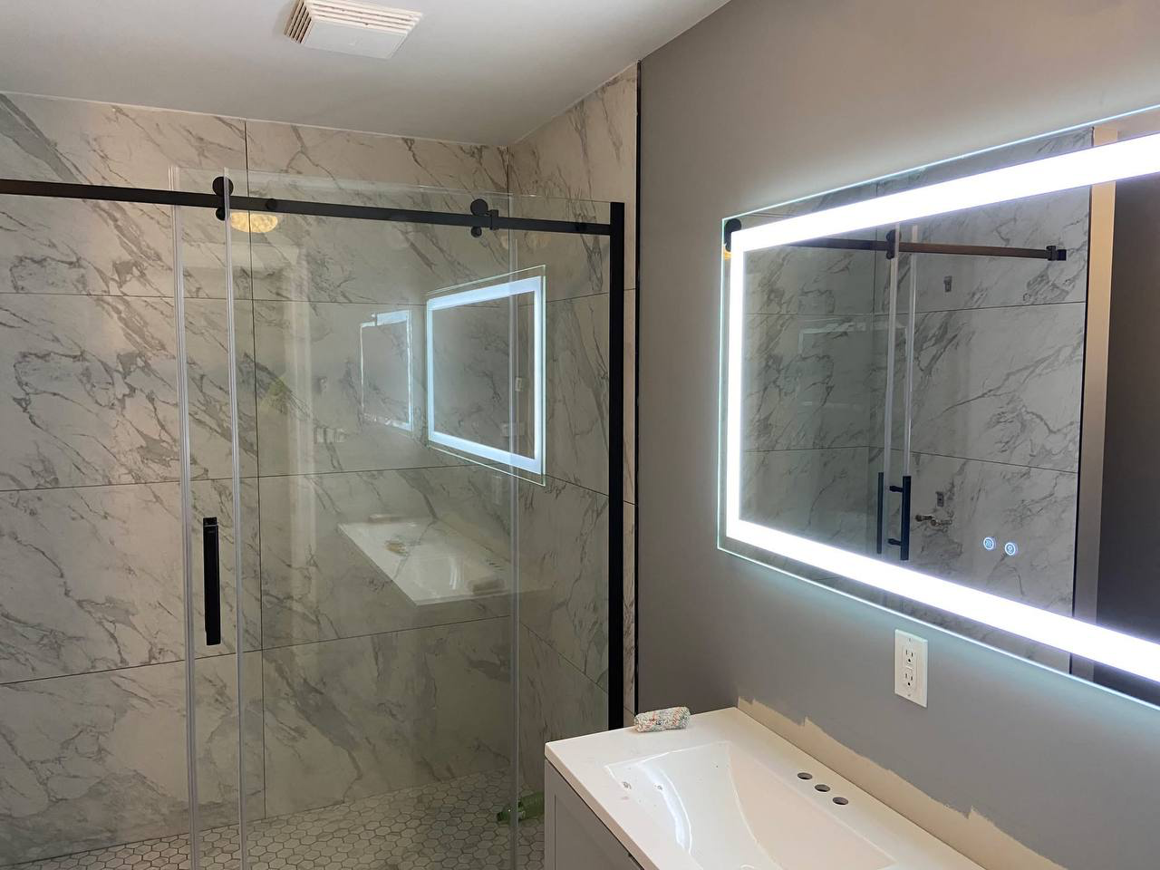 A modern bathroom featuring a glass-enclosed shower with marble walls and a large illuminated mirror above a white sink.