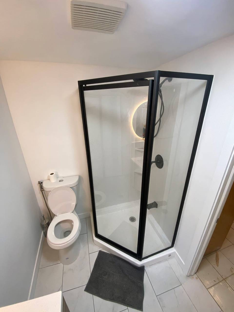 Bathroom with a toilet on the left and a glass shower stall on the right, with a circular mirror and sink visible through the shower glass, and a gray bath mat on the tiled floor.