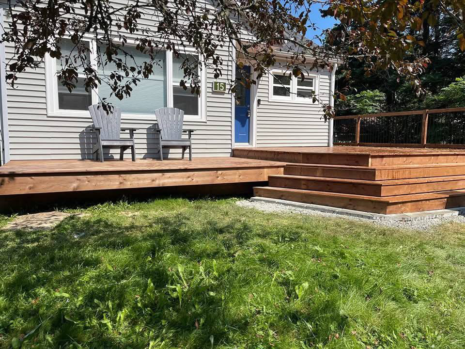 Front porch of a house with wooden stairs and deck, two gray Adirondack chairs, house with gray siding, blue door, house number 15, and a tree with brown and green leaves in the foreground.