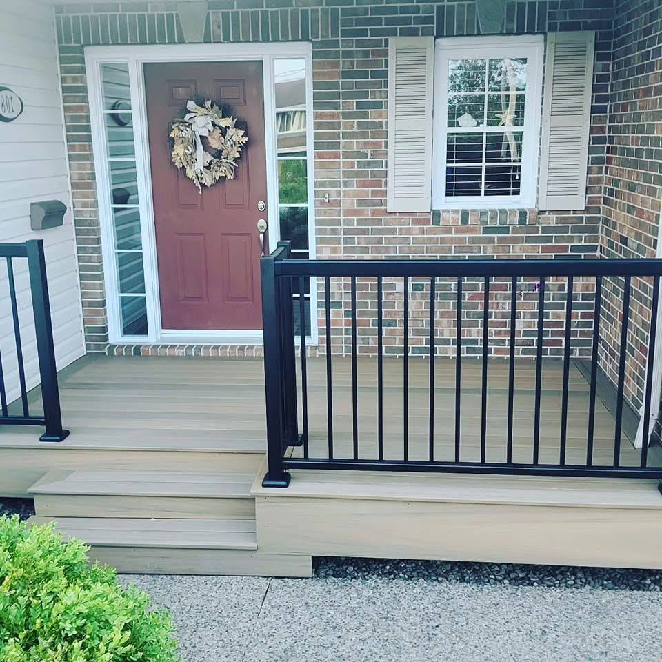 Front porch with a brown door decorated with a wreath, a small set of steps leading to a covered porch, black metal railing around the porch, brick and siding exterior walls, a window with white shutters, and a small shrub in front.
