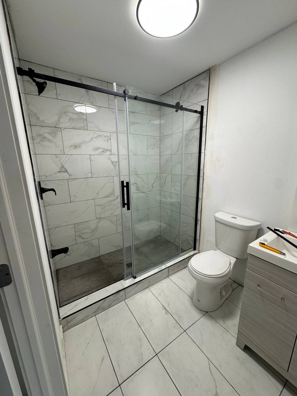 Modern bathroom with a clear glass shower enclosure, white marble tiles, and a white toilet next to a light wood vanity with tools on top.