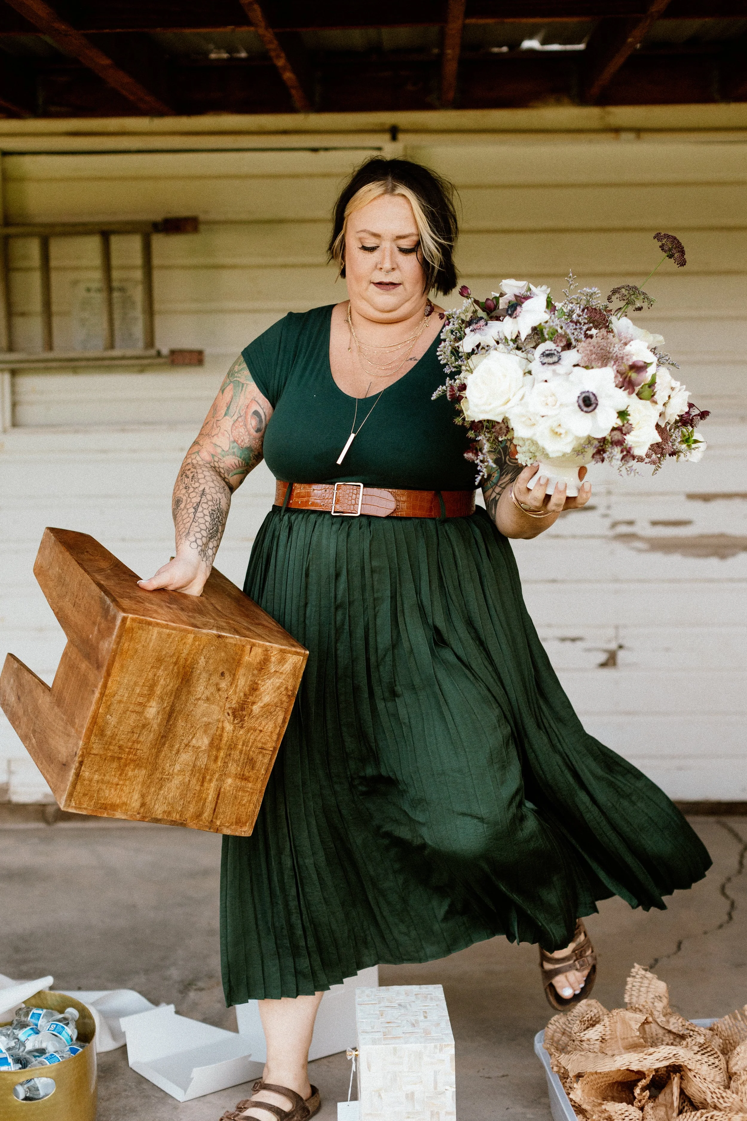 Woman with tattoos wearing a green dress, holding a bouquet of flowers and a wooden block, standing in front of a beige wooden wall.