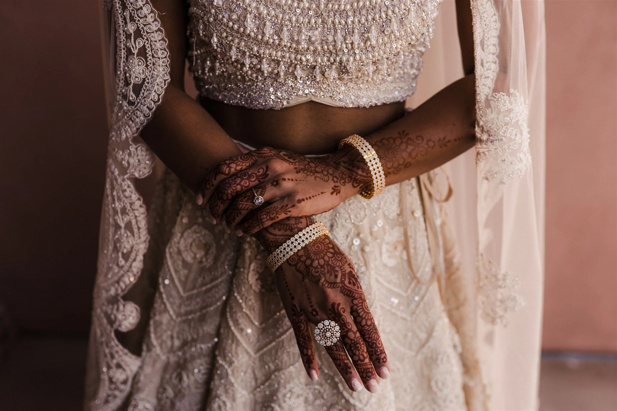 A woman wearing a heavily embellished beige blouse with lace and beaded details, with her hands clasped showing henna tattoos, wearing diamond jewelry including a ring, bracelet, and ring, in front of a matching lace skirt and sheer lace dupatta.