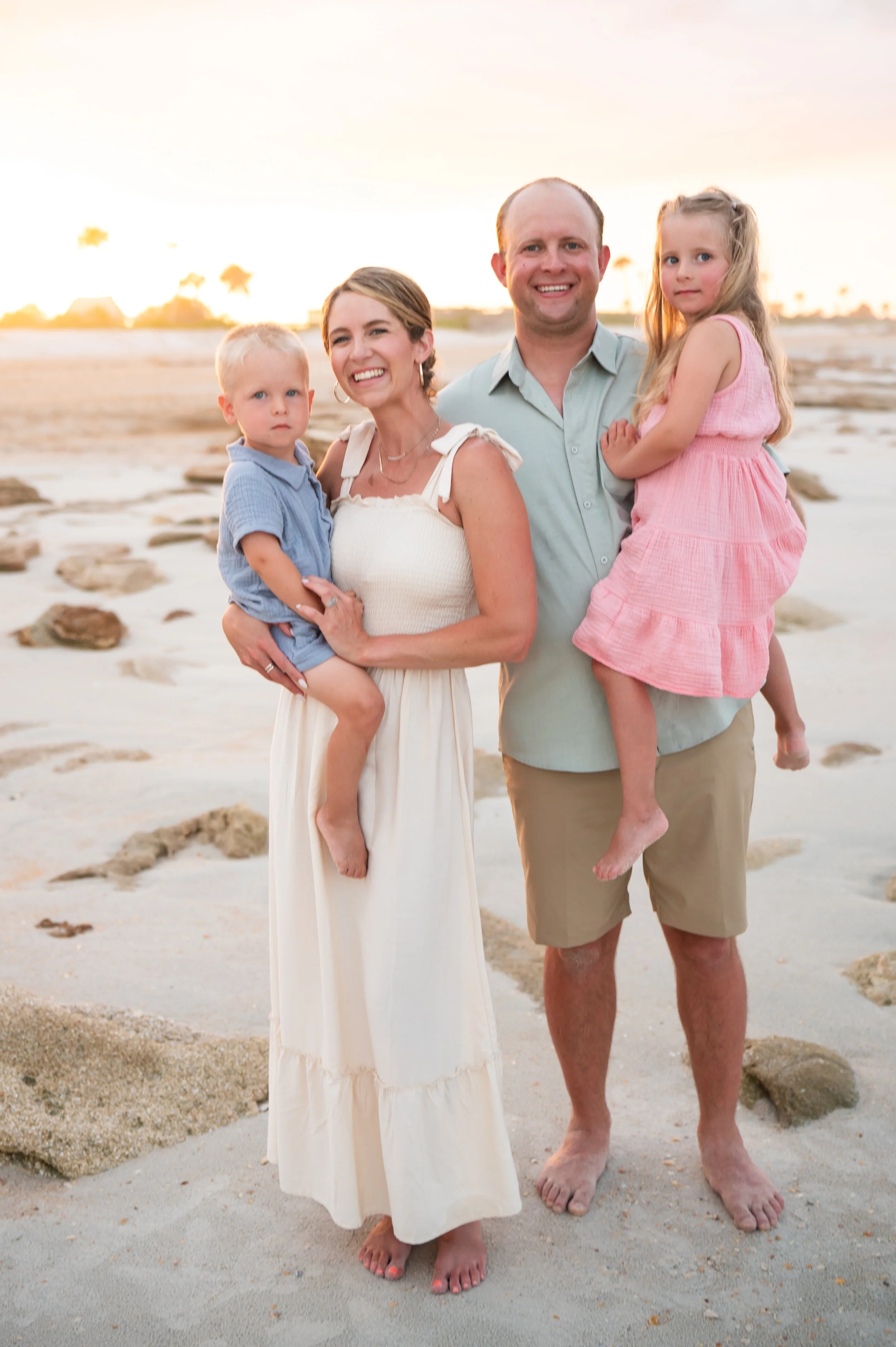 A smiling family of four standing on a beach at sunset. The mother is holding a young boy, and the father is holding a young girl. The beach has rocks scattered across the sand.