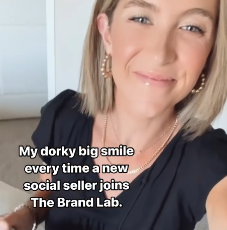 A woman with short blonde hair smiling in a close-up selfie. She is wearing hoop earrings, a gold necklace, and a black top. There is text overlay that says: "My dorky big smile every time a new social seller joins The Brand Lab."