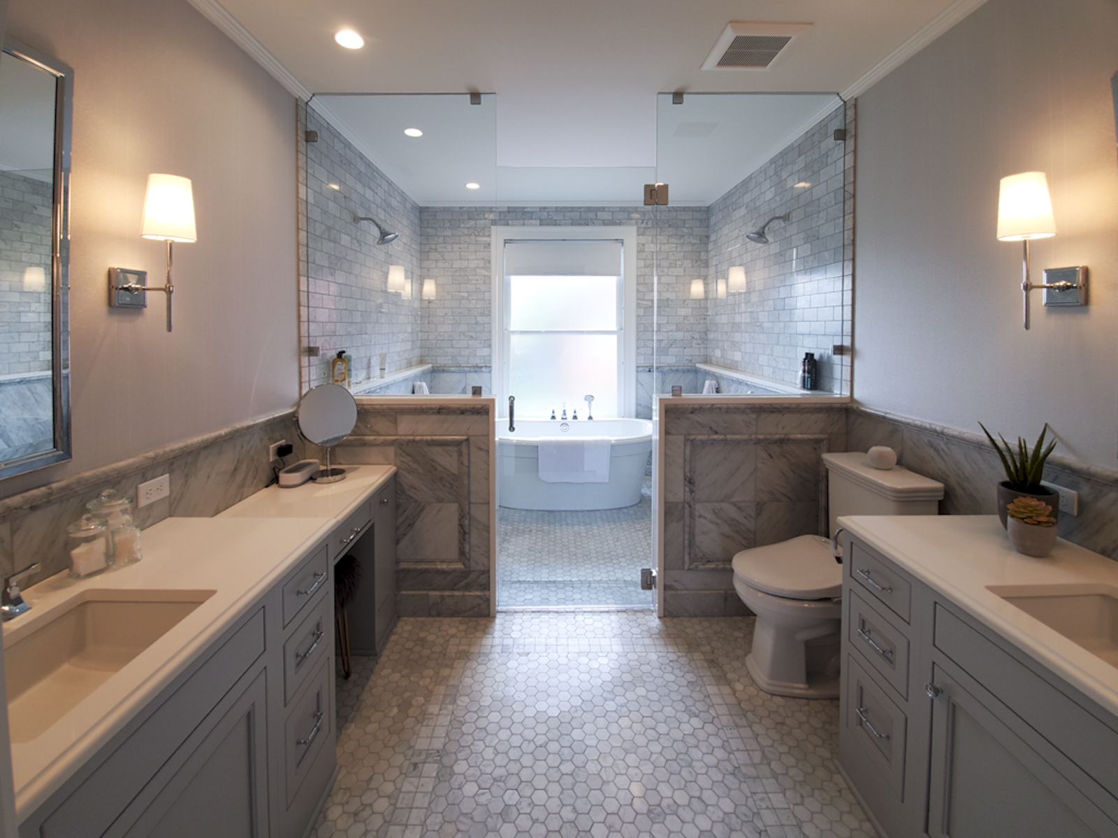 A spacious bathroom with double vanities, a toilet, and a freestanding bathtub in a separate space with a frosted window. The room features light-colored walls, marble countertops, and hexagonal tile flooring.