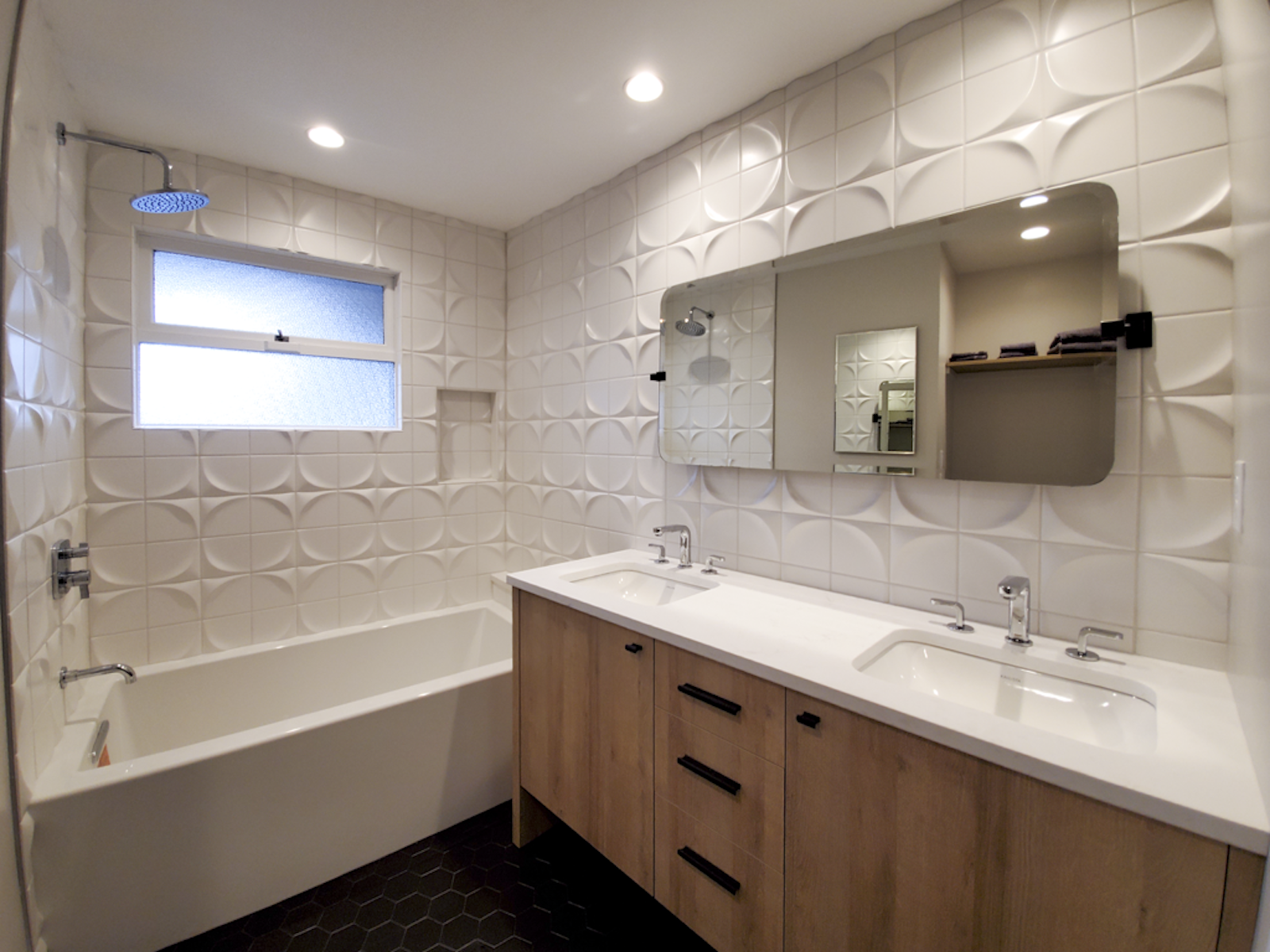 Modern bathroom with white textured wall tiles, a frosted window, a double vanity with a white countertop, two sinks, and a wall-mounted mirror. There is a bathtub with wall-mounted faucets and a showerhead in the corner.