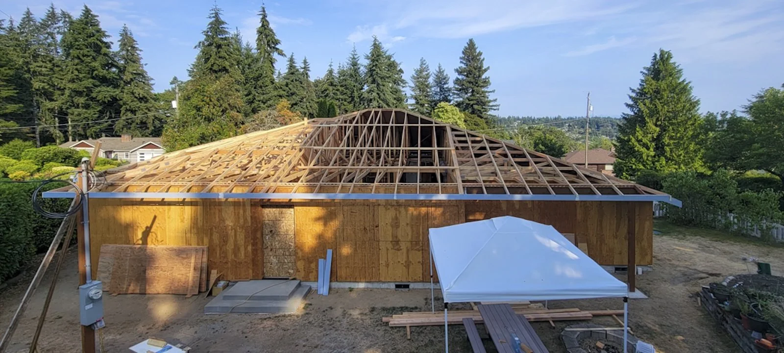 House under construction with wooden framework and partial walls, surrounded by trees and construction equipment.