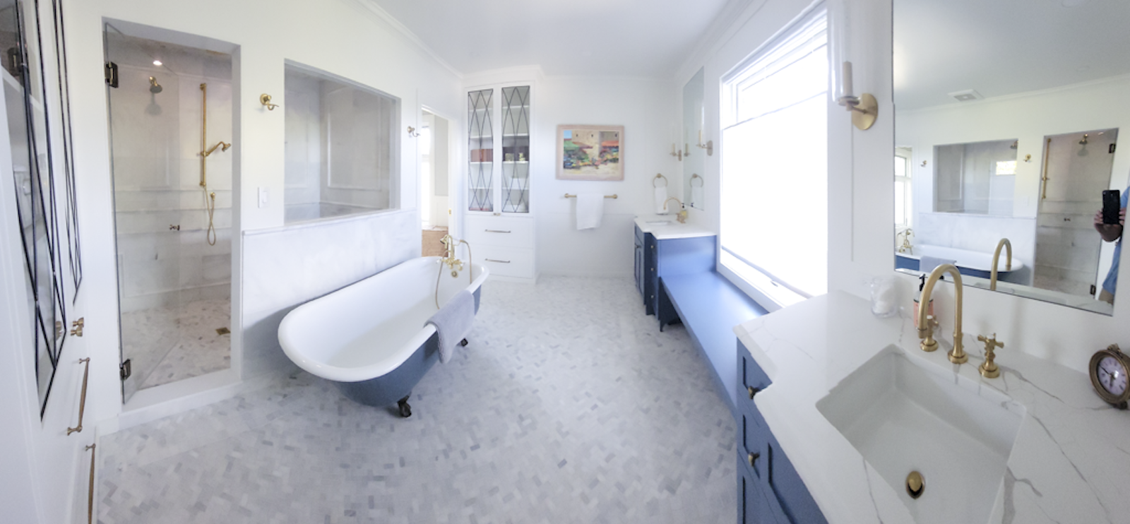 A spacious bathroom with a separate shower, a clawfoot bathtub, a blue vanity, gold fixtures, and a large window with natural light.