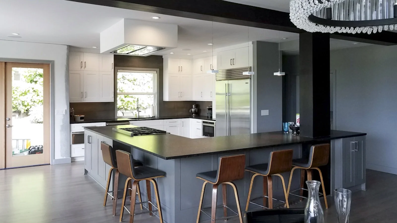 Modern kitchen with white cabinets, black countertops, a large central island with a stove, brown chairs, stainless steel refrigerator, and a chandelier overhead.