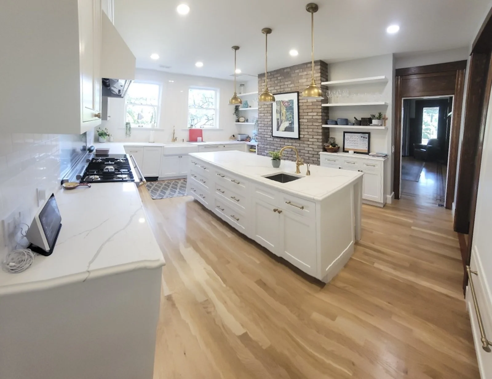 Bright, modern kitchen with white cabinetry, a large central island with a sink, and three hanging gold pendant lights. Wooden floors, brick accent wall, and open shelving with dishes and decor. Windows let in natural light.