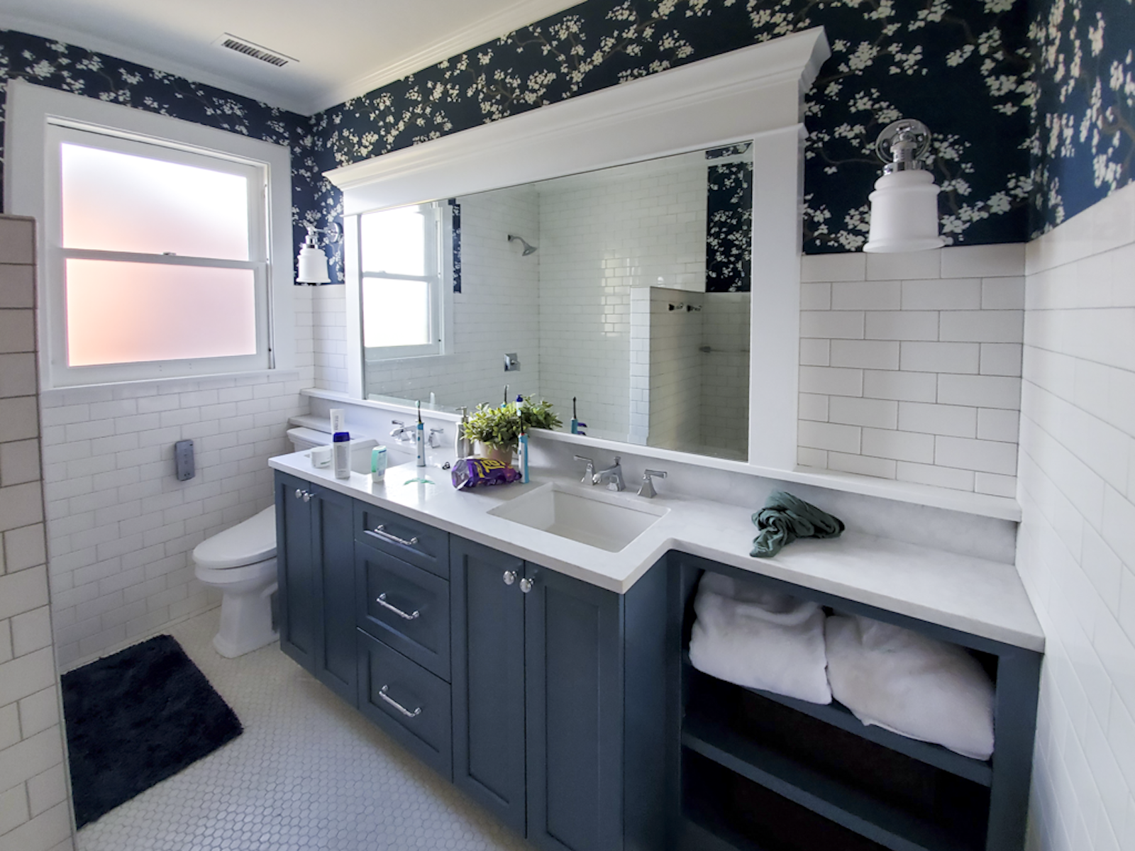 Modern bathroom with blue vanity, white countertop, large mirror, white subway tile walls, floral wallpaper above, two windows, black rug, towels, toiletries, and decorated with plants.