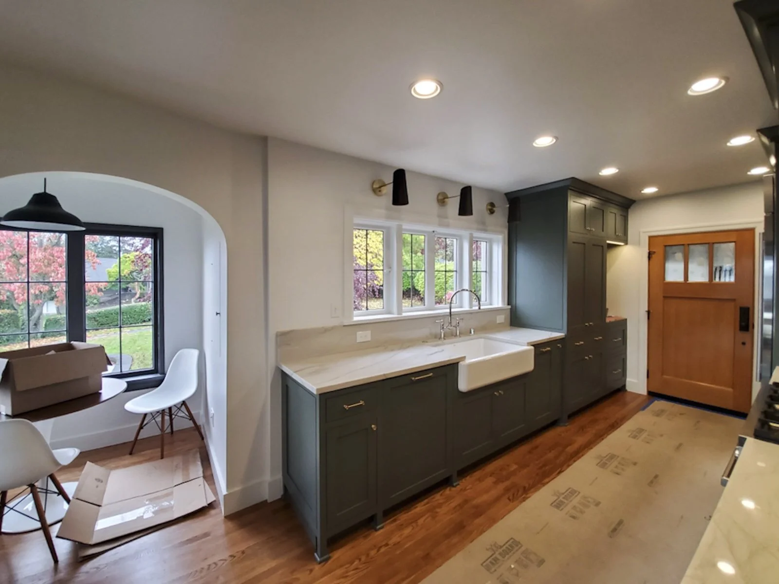 Kitchen with dark green cabinets, white farmhouse sink, wooden door, hardwood floors, and small dining area with window showing trees outside.
