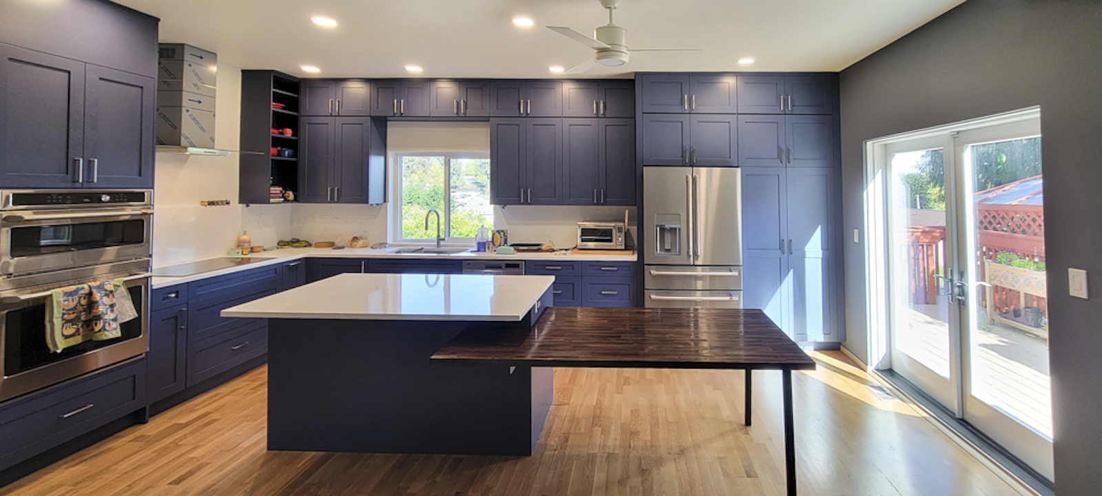 Modern kitchen with blue cabinets, stainless steel appliances, white countertops, a kitchen island, hardwood floors, and sliding glass doors leading outside.