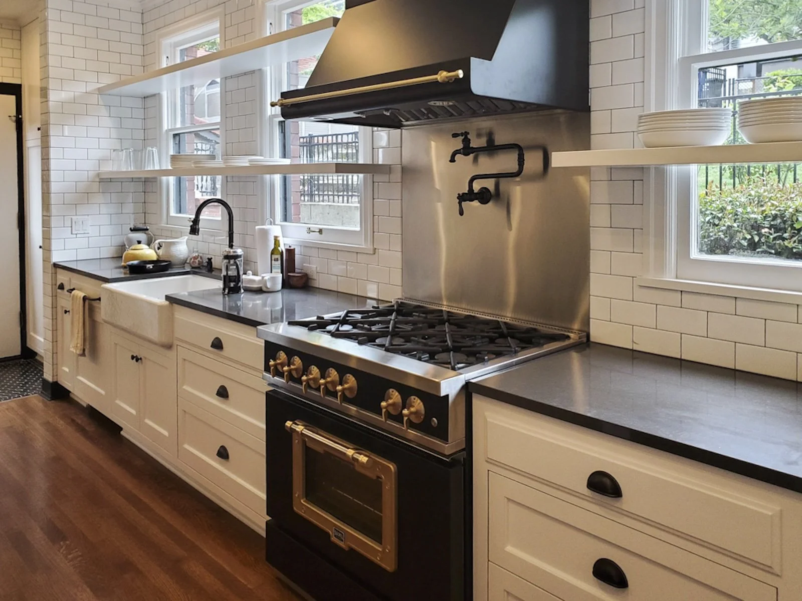 Modern kitchen with white cabinetry, black countertops, a farmhouse sink, open shelves with plates and glasses, a stainless steel stove with gold handles, and windows with black iron grills overlooking greenery.