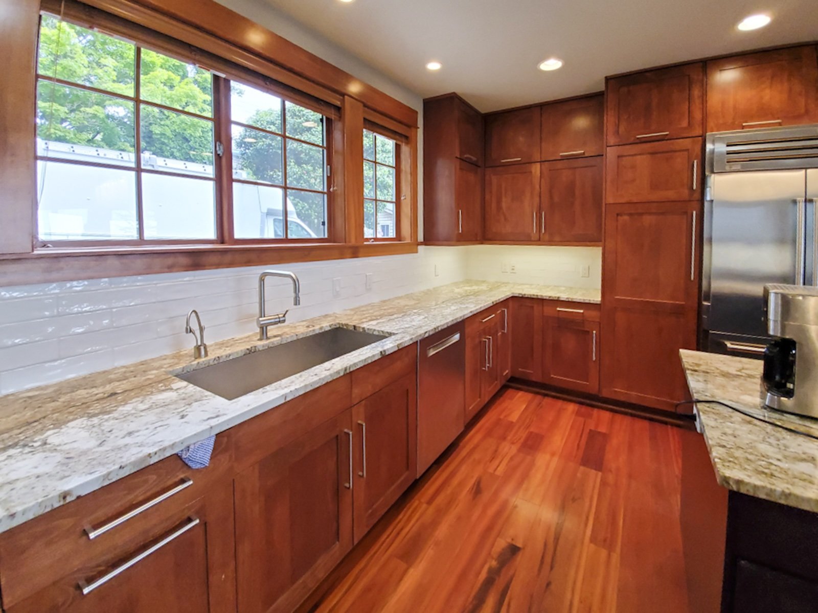 A kitchen with wooden cabinets, granite countertops, a double window over the sink, hardwood floors, and stainless steel appliances.