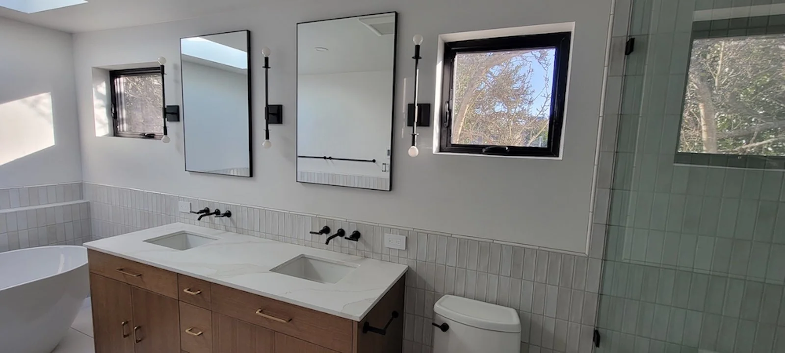 Modern bathroom with white walls, two small windows, a wooden vanity with two sinks, black fixtures, two mirrors, and black wall-mounted lights.