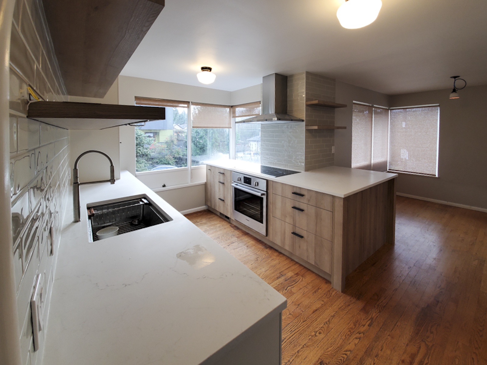 Modern kitchen with wooden cabinets, white countertops, a stainless steel oven, large windows with shades, and wooden flooring.