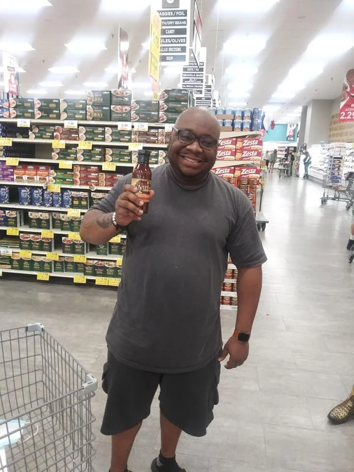 A man standing in a grocery store aisle, smiling and holding a bottle of sauce or condiment.