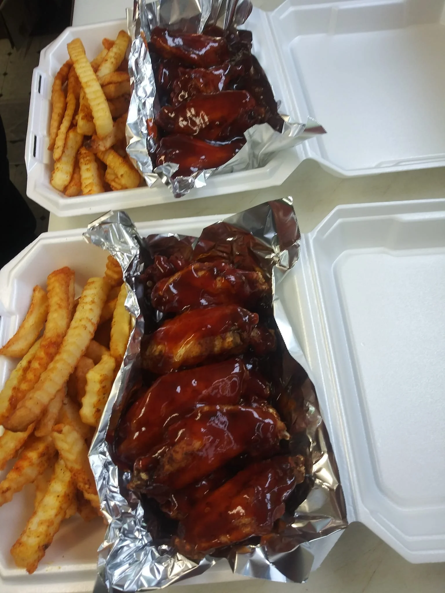 Two takeout containers with crinkle-cut French fries and servings of barbecue chicken wings covered in barbecue sauce.